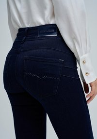Primer plano de una persona que lleva puesto un pantalón vaquero de mezclilla azul oscuro con bolsillos traseros decorados con pequeñas cuentas, combinado con una camisa blanca de manga larga.