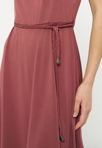 Πλεξούδα λεπτή ζώνη με μεταλλικές άκρες δεμένη γύρω από τη μέση φορέματος χωρίς μανίκια σε στυλ σκόνης ροζ, που δείχνει με το δεξί χέρι να ακουμπά δίπλα στο σώμα.