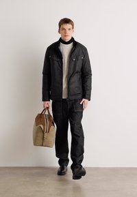 Schwarze isolierte Jacke über einem hellgrauen Pullover, kombiniert mit dunklen Jeans. Trägt eine braun-weiße Reisetasche und schwarze Schuhe.