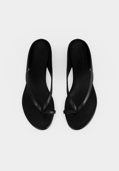 Ζευγάρι μαύρα μινιμαλιστικά σανδάλια στυλ στρινγκ με λεπτές λουρίδες και επίπεδες σόλες, απεικονίζονται σε λευκό φόντο.