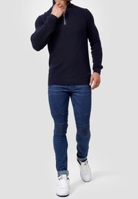 Granatowy sweter z prążkowanego dzianiny z kołnierzem na ćwiartkę, zestawiony z niebieskimi dżinsami z prążkowanymi akcentami i białymi trampkami.