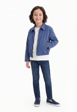 Παιδί με καστανά μαλλιά στέκεται χαμογελώντας, φοράει μια μπλε ζακέτα με φερμουάρ, λευκό πουκάμισο, σκούρο τζιν και ναυτικά παπούτσια χωρίς κορδόνια, σε λευκό φόντο.