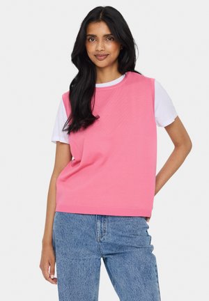 Pulover roz fără mâneci, cu un model texturat, cu tiv striat, purtat peste un tricou alb; asortat cu blugi albaștri deschiși.