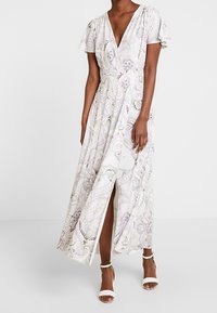 Maxi dress in light fabric featuring a paisley pattern, V-neck, flutter sleeves, and a side slit, complemented by white strappy heels.