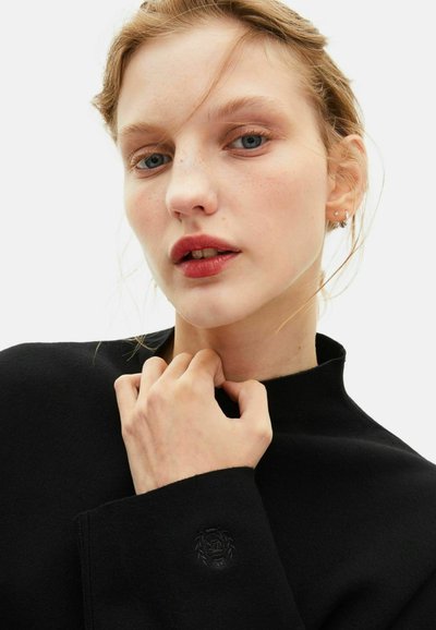 Giovane donna con pelle chiara e labbra rosse che indossa un maglione nero a collo alto, tenendo il colletto vicino al mento, con ciocche di capelli sciolti.