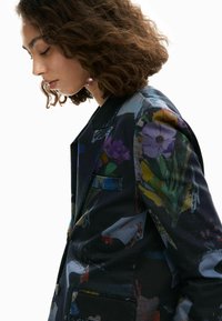 Blazer nero con stampa floreale dai colori vivaci. Realizzato in tessuto liscio, presenta un rever a incastro e tasche laterali. Silhouette aderente.