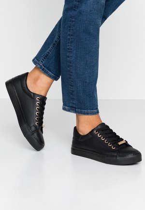 Baskets basses en cuir noir avec lacets noirs et œillets dorés, portées avec un jean en denim bleu sur un sol blanc.