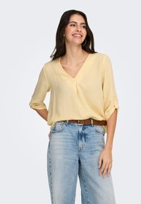 Geel, lichtgewicht blouse met V-hals en driekwart opgerolde mouwen, gecombineerd met lichtblauwe, losse jeans en een bruine riem.