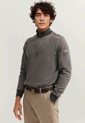 Młody mężczyzna z kręconymi włosami, ubrany w szary sweter z zamkiem do ćwiartki i beżowe spodnie, stoi z jedną ręką w kieszeni na jednolitym tle.