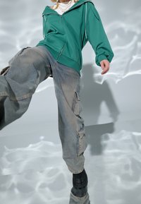 Sudadera verde con cremallera y acentos blancos, combinada con unos pantalones cargo sueltos de color azul claro con múltiples bolsillos y una textura descolorida. Botas negras.
