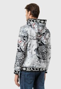 Haut à capuche gris avec des imprimés graphiques noirs, présentant un design de crâne et des motifs complexes. Les poignets en tricot noir et l'ourlet apportent un contraste.