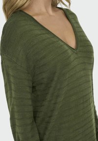 Pull en tricot vert olive avec col en V, présentant de fines rayures verticales, coupe ample et tissu texturé. Matière douce et légère.
