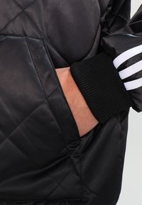 Chaqueta acolchada negra con un bolsillo lateral y puño negro acanalado. Tres rayas blancas acentúan la manga. Textura suave y brillante.