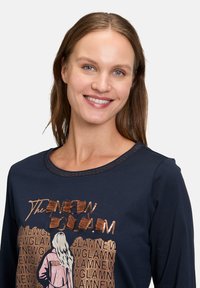 Marineblaues Langarmshirt mit rundem Ausschnitt, verziert mit metallischen Akzenten und einem grafischen Druck einer Frau mit dem Text "The New Glam".