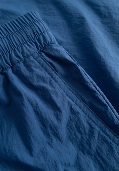 Blauer Stoff mit einer knitterigen Textur, einer elastischen Taille und einer Seitentasche. Licht spiegelt sich auf der unebenen Oberfläche.