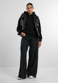 Gilet imbottito nero sopra un hoodie nero, abbinato a pantaloni cargo neri a gamba larga. L'outfit presenta una vestibilità rilassata e materiali testurizzati.