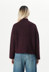 Person med langt blondt hår iført en mørk lilla genser og blå jeans, stående med ryggen til mot en hvit bakgrunn.