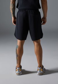 Svarte treningsshorts med elastisk liv, glatt tekstur og sidelomme, kombinert med grå og gull løpesko.