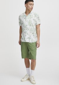 Camisa blanca de manga corta con un patrón floral verde, combinada con pantalones cortos verdes hasta la rodilla, calcetines blancos y zapatillas de deporte beige.
