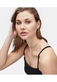 Schwarzes Tanktop mit dünnen Trägern, kombiniert mit einer goldenen Kettenhalskette und kleinen goldenen Creolen. Glatte Hauttextur und dezentes Make-up.