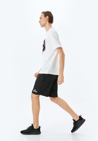 Hvid t-shirt med et grafisk design, sorte shorts og sorte træningssko. Model går med en neutral baggrund.