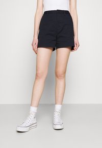 Pantalones cortos de un azul marino oscuro con cintura alta, con un diseño ajustado, bolsillos laterales y una textura de algodón suave. Combinados con zapatillas blancas.