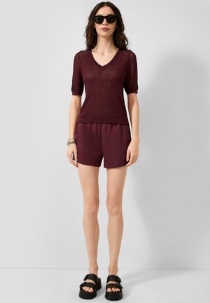 Femme portant un haut en maille bordeaux foncé à manches courtes et un short assorti, des sandales noires, des lunettes de soleil et des bracelets, debout devant un fond uni.