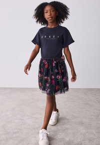 Marineblaues T-Shirt mit kurzen Ärmeln und goldenen Buchstaben, kombiniert mit einem schwarzen Tüllrock mit Blumenmuster. Dazu weiße Sneaker mit einer Schleifenakzentuierung.