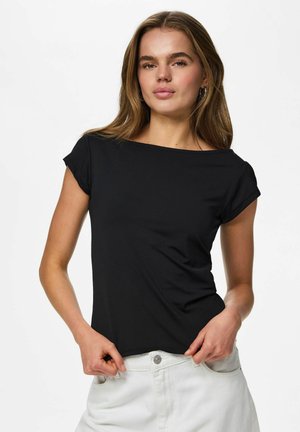 Ung kvinde med langt hår iført en sort top med korte ærmer og hvide bukser, holder i kanten af sin top, neutral baggrund.