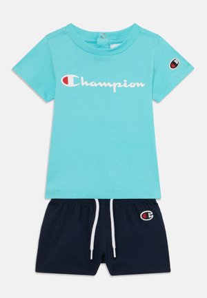 Modra kratka majica s kratkimi rokavi z belim logotipom "Champion", v kombinaciji s črnimi kratkimi hlačami, ki imajo izvezen logotip in belo vrvico.