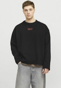 Felpa nera oversized con spalle cadute, caratterizzata da un logo ricamato rosso sul petto. Abbinata a jeans azzurri chiari.