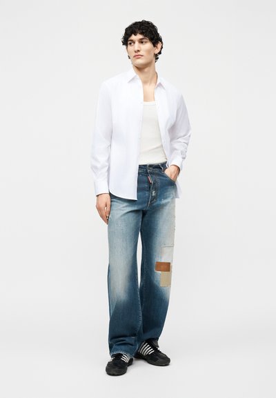 Jeune homme debout portant une chemise blanche à boutons sur un débardeur blanc, un jean large bleu avec des morceaux réparés et des baskets noires à rayures blanches.