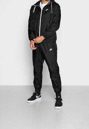 Mand iført sort Nike-træningsdragt med hvide logoer og sorte Nike-sneakers, stående foran en ensfarvet lys baggrund.