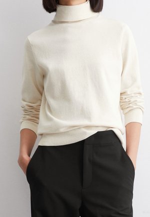 Femme portant un pull à col roulé crème avec les manches remontées, légèrement rentré dans un pantalon noir taille haute, les mains dans les poches.