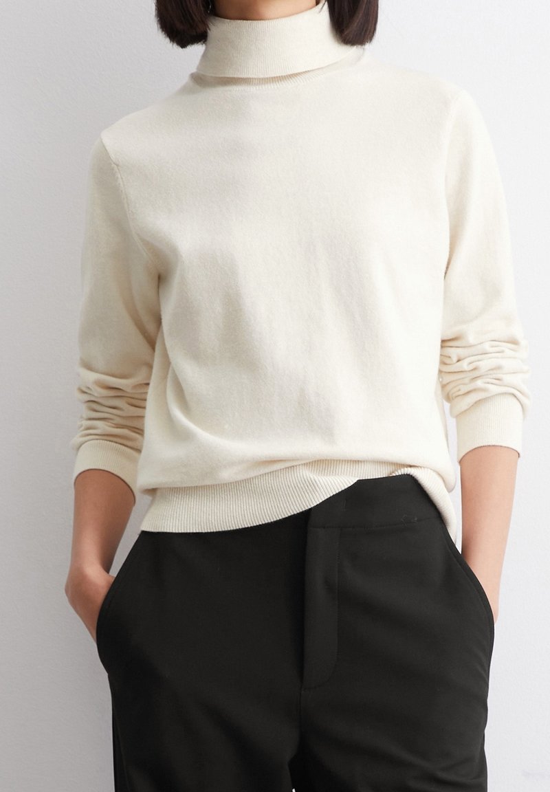Femme portant un pull à col roulé crème avec les manches remontées, légèrement rentré dans un pantalon noir taille haute, les mains dans les poches.
