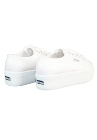 Baskets en toile blanches avec une texture lisse, à lacets, et une semelle compensée blanche surélevée. Présente un petit logo noir sur le talon.