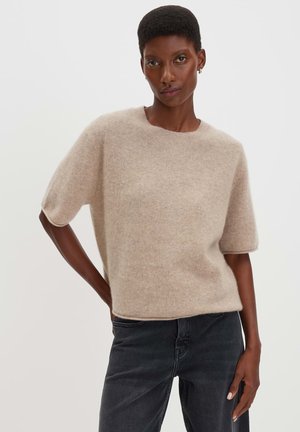 Dark-skinned person wearing a beige short-sleeve fuzzy sweater and black jeans, standing with one hand on hip against a plain light background.