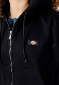 Czarna hoodie zapinana na zamek, wykonana z mieszanki bawełny, z przednią kieszenią, kapturem ze sznurkiem i metką na piersi. Gładka tekstura.