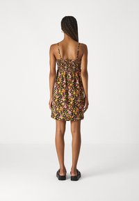 Frau mit geflochtenem Haar, die ein ärmelloses Kleid mit Blumenmuster und schwarze Plateausandalen trägt, steht mit dem Rücken zu einer weißen Hintergrund.