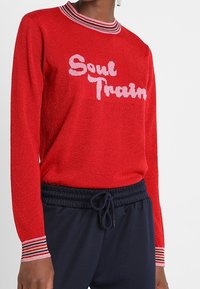 Rotes Langarmoberteil mit glitzernder Textur, versehen mit dem Schriftzug "Soul Train" in Weiß, und gestreiftem geripptem Rundhalsausschnitt sowie Bündchen.