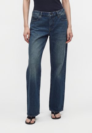 Personne portant un jean droit bleu foncé, des sandales noires à bout ouvert et un haut noir sans manches, debout devant un fond blanc uni.