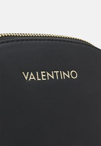 Černá kožená taška se zlatým zipem. Na přední straně je vytlačené logo "VALENTINO". Hladký povrch, strukturovaný tvar.