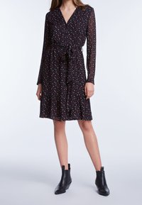 Zwarte jurk met lange mouwen en een V-hals, voorzien van een strik taille, gerimpelde zoom en een klein, meerkleurig bloemenpatroon. Gecombineerd met zwarte enkellaarsjes.