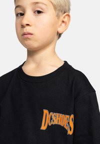 Černé bavlněné tričko s výrazným oranžovým logem "DCSHOES" na levé části hrudi. Klasický kulatý výstřih a krátké rukávy.