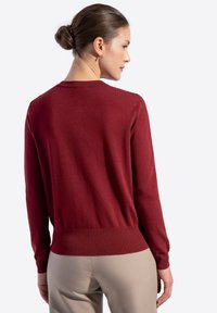 Maglione bordeaux con maniche lunghe, orlo a coste e colletto rotondo. Texture liscia, design aderente, mostrato di spalle con pantaloni di colore neutro.