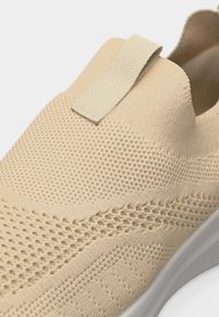 Μπεζ πλεκτό αθλητικό παπούτσι slip-on με υφασμάτινη, διαπνέουσα υφή στην επάνω επιφάνεια και υφασμάτινη θηλιά στο ελαστικό κολάρο, λευκή εξωτερική σόλα εν μέρει ορατή.