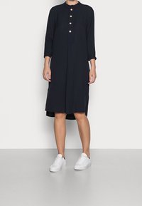 Abito nero al ginocchio con colletto, tre bottoni davanti e maniche di lunghezza media; abbinato a sneaker bianche. Tessuto di texture liscia.