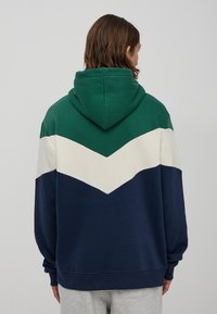 Kaptur przeciwsłoneczny z miękkiego materiału z zielonym, kremowym i granatowym pasem w wzór chevron na plecach. Luźny krój z kieszenią kangurką.