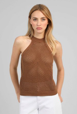 Femme aux longs cheveux blonds portant un haut marron en crochet sans manches et un pantalon blanc, debout devant un fond gris uni.