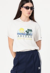 Balta medvilninė marškinėlių su spalvinga grafika, kuri vaizduoja saulę, palmės ir tekstą „ADIDAS Sporto Klubas“. Pora su tamsiai mėlynomis šortais.